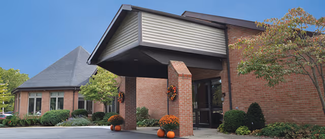 Exterior view of a brick assisted living facility with a covered entrance, decorated with fall-themed pumpkins and flowers, surrounded by green shrubs and trees under a clear blue sky.