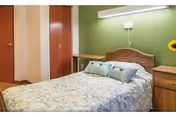 A bedroom with a single bed covered in a floral bedspread and two pillows. The bed has a wooden headboard and is positioned against a green wall with a mounted light fixture above. There is a wooden nightstand with a sunflower in a vase on the right side of the bed. Two closed wooden doors are visible in the background.