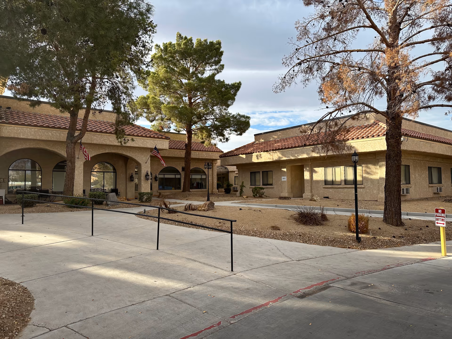 Exterior view of Mission Pines Nursing & Rehabilitation Center showing a beige stucco building with red tile roofing, arched windows, and two American flags. There are trees and a concrete walkway with a black handrail leading to the entrance. A 'No Parking Anytime' sign is visible near the driveway.