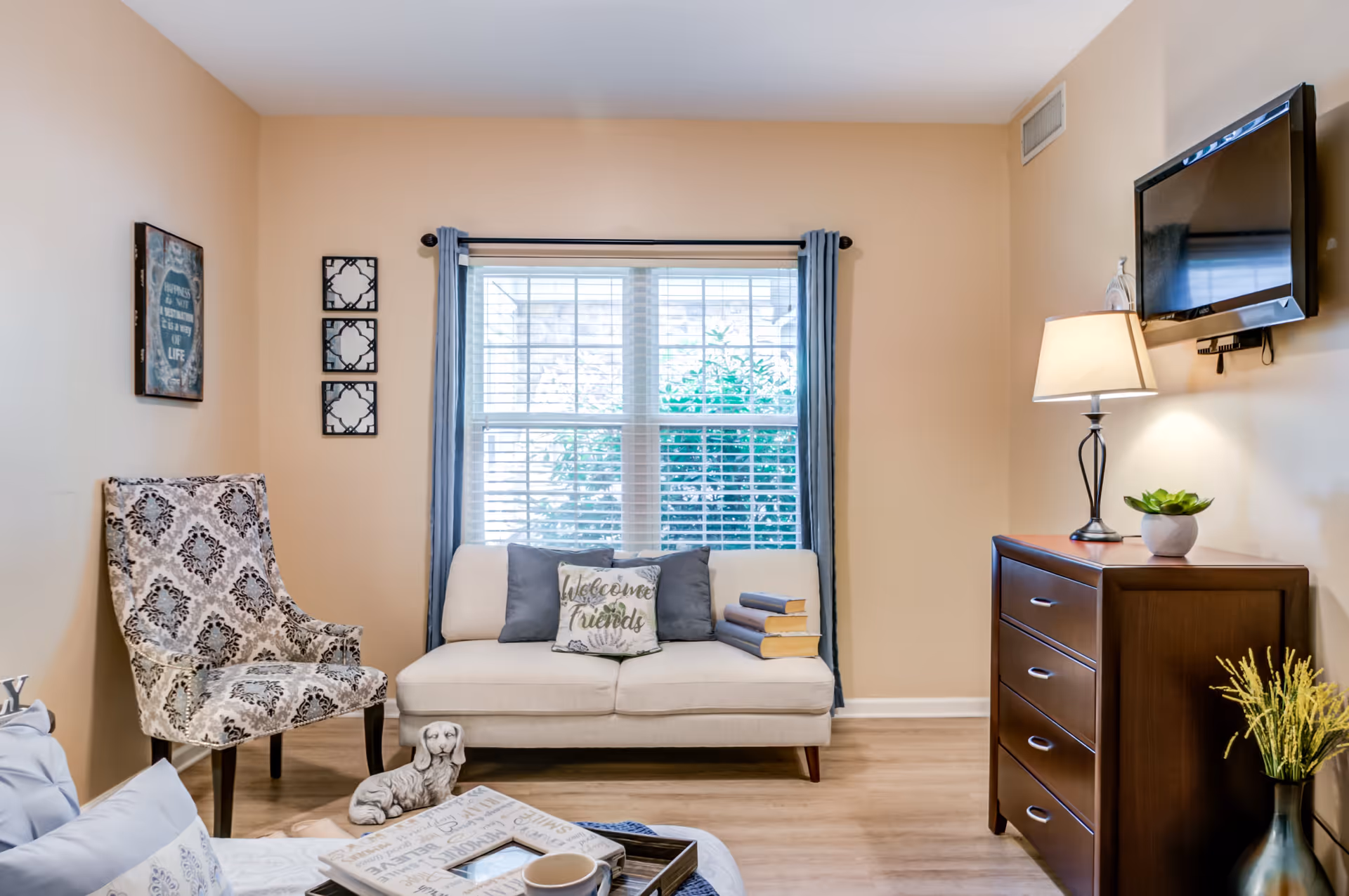 A cozy living room with a beige loveseat adorned with three pillows, one of which says 'Welcome Friends'. Next to the loveseat is a patterned armchair. A wooden dresser with a lamp, a small plant, and a wall-mounted TV is on the right. The room has a large window with blue curtains and light-colored walls and flooring.