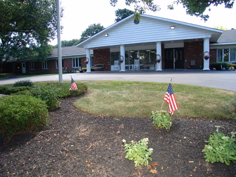 Front exterior view of a single-story brick building with a white triangular roof entrance supported by columns. There are two American flags planted in the garden area in front of the building, which has some shrubs and small plants. The driveway leads up to the entrance with glass doors and windows.
