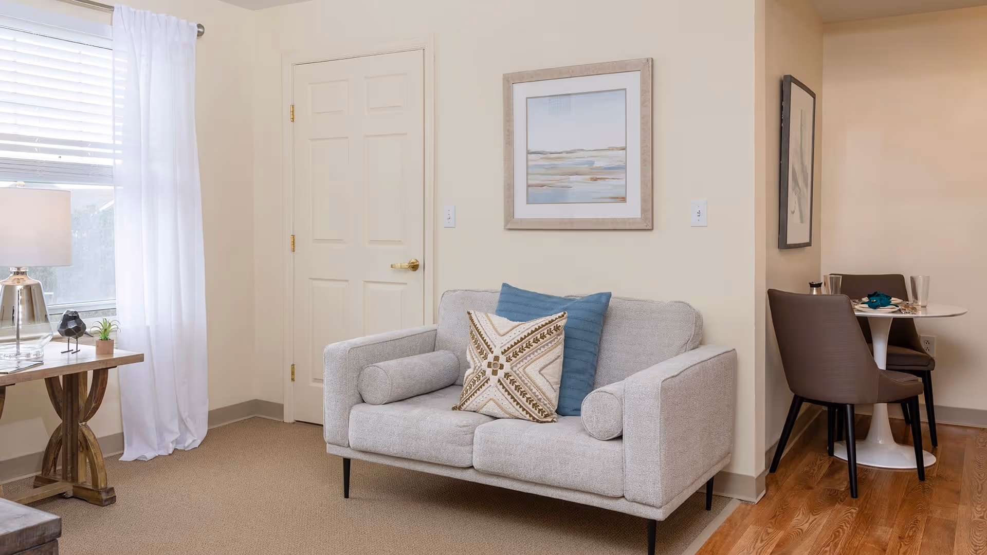 A cozy living room area with a light gray loveseat adorned with two decorative pillows, one blue and one patterned. To the left, there is a wooden side table with a lamp, a small plant, and a decorative bird figurine. A window with white curtains allows natural light to fill the room. On the right side, a small dining area with a round white table and two brown chairs is partially visible. The walls are painted beige and decorated with framed artwork.