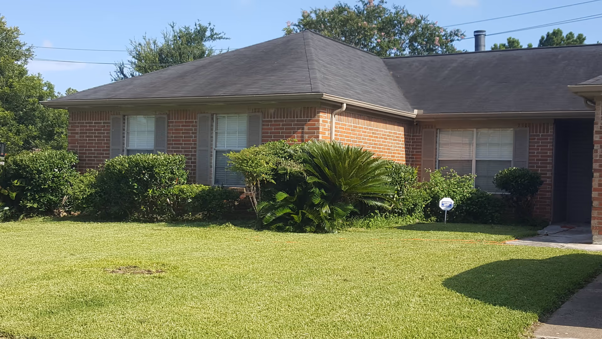 Front exterior view of a single-story brick building with a dark shingled roof, surrounded by green bushes and a well-maintained lawn under a clear blue sky.