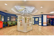 Interior view of a senior living facility lobby area with a central white display case under a skylight, blue walls, seating area with green chairs, and a grocery store section visible on the right.