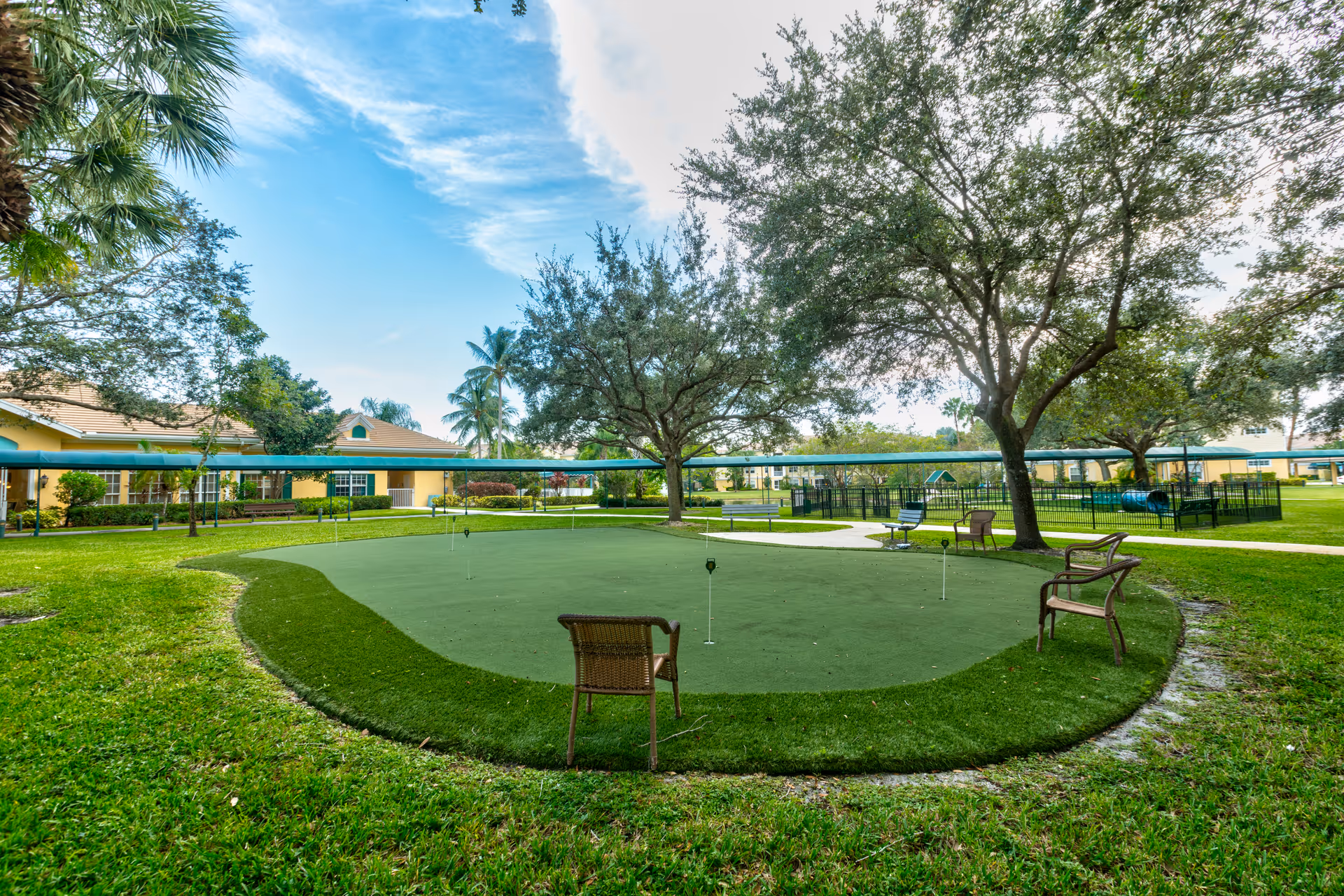 Outdoor area at Aston Gardens At Parkland Commons featuring a putting green with several small flags, surrounded by green grass and trees. There are a few chairs placed around the putting green, and buildings are visible in the background under a partly cloudy blue sky.
