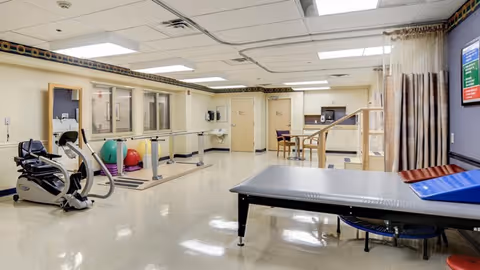 A rehabilitation room in Easton Skilled Nursing and Rehabilitation Center featuring exercise equipment including a stationary bike, parallel bars for walking practice, colorful exercise balls, a padded therapy table, and a small table with chairs. The room has a clean, bright interior with tiled floors and fluorescent lighting.