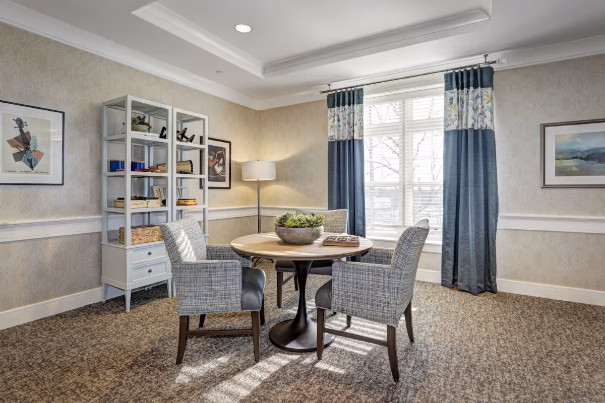 A cozy interior room with a round wooden table surrounded by four upholstered chairs. A decorative plant centerpiece is on the table. The room has beige patterned wallpaper, a carpeted floor, a tall floor lamp in the corner, a white shelving unit with books and decor, and large windows with blue and floral curtains letting in natural light. Framed artwork hangs on the walls.