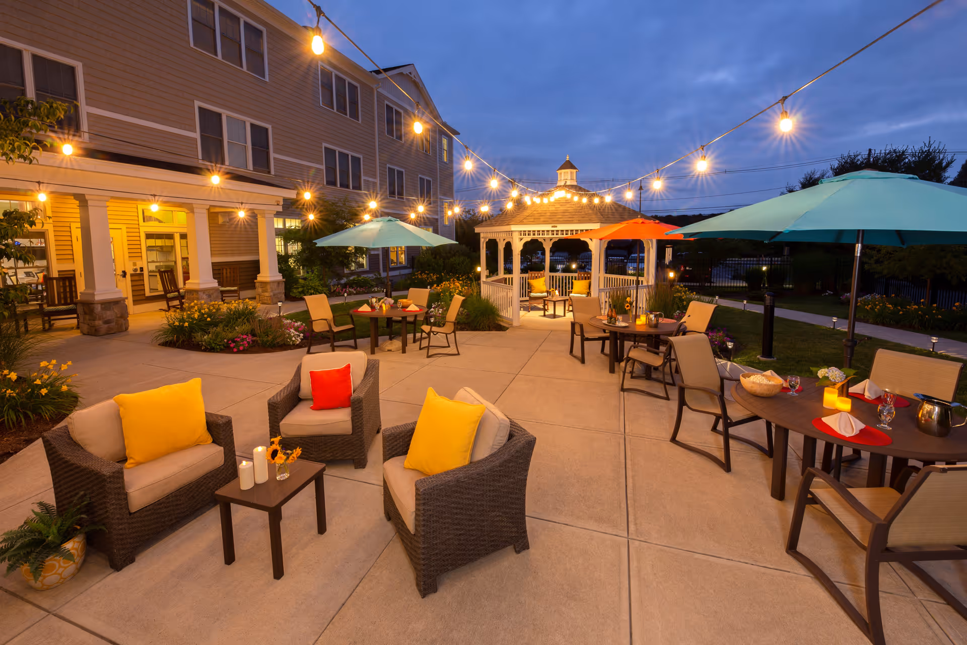 Outdoor patio area at dusk with string lights hanging overhead, several tables with umbrellas, wicker chairs with colorful cushions, a small gazebo in the background, and a building with lit windows on the left side.