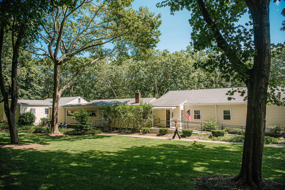 A single-story yellow building surrounded by trees and greenery under a clear blue sky. A person is walking on the sidewalk in front of the building, which has an American flag near the entrance and a wheelchair ramp. The area is well-maintained with grass and shrubs.