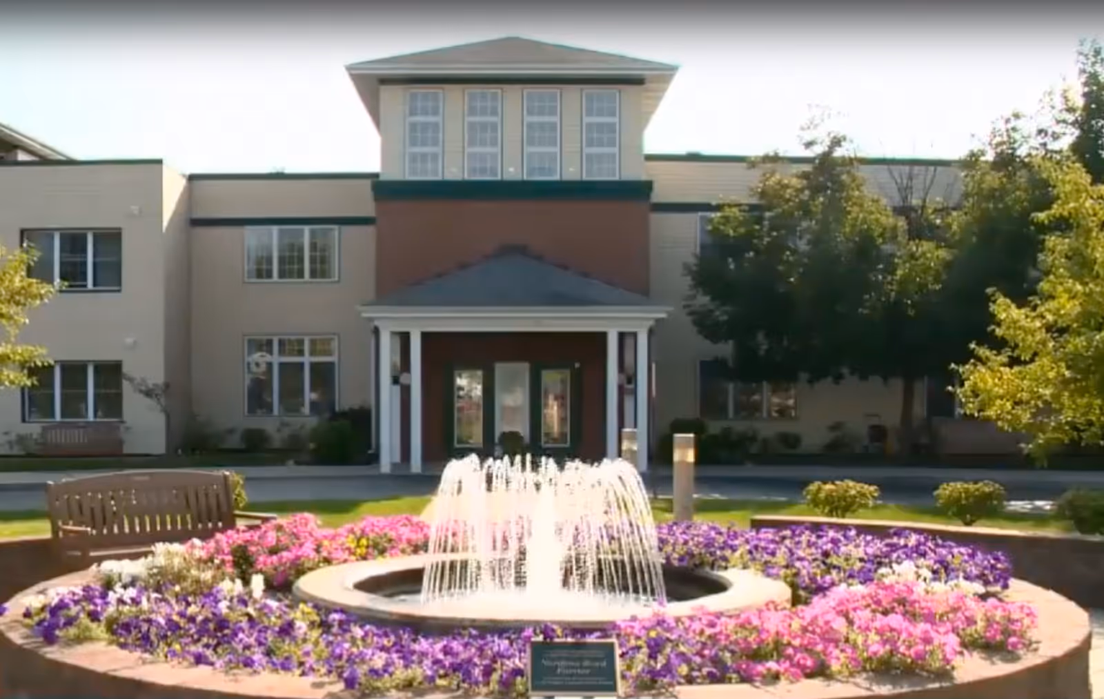 Front exterior view of GreenField Terrace facility with a circular flower bed featuring purple, pink, and white flowers surrounding a water fountain. There are benches around the flower bed and trees on either side of the building entrance.