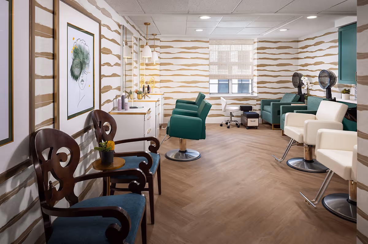 Interior view of a salon area in a senior living facility featuring multiple salon chairs in green and white, hair dryers, a small seating area with two wooden chairs and a small table, and decorative wall art. The room has patterned wallpaper and a window with a woven shade.