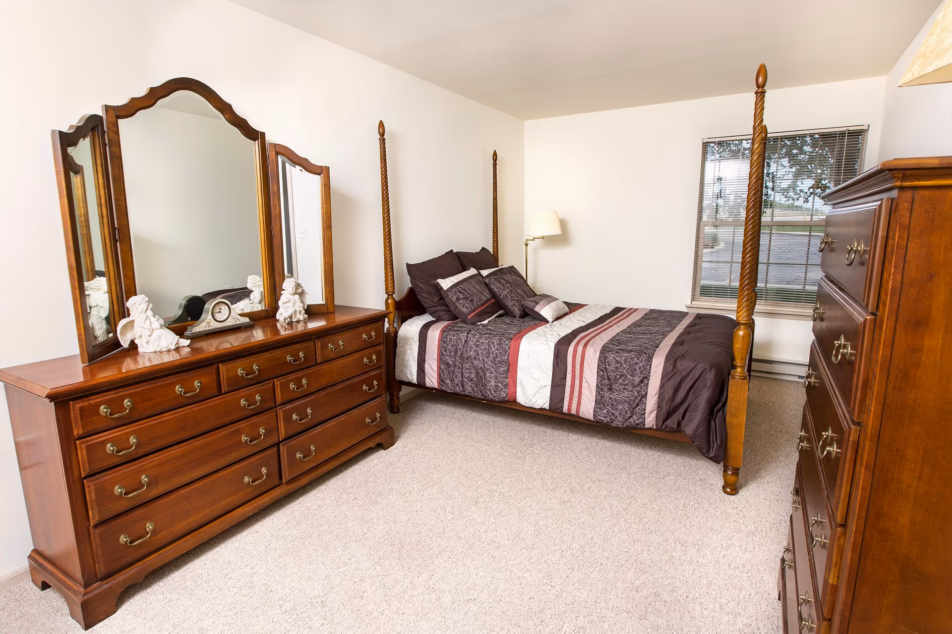A bedroom with a wooden four-poster bed dressed in a striped comforter and multiple pillows. There is a wooden dresser with a large three-panel mirror on the left side, decorated with two angel figurines and a clock. On the right side, there is a tall wooden chest of drawers. A window with blinds is visible on the far wall, and a floor lamp stands next to the bed.