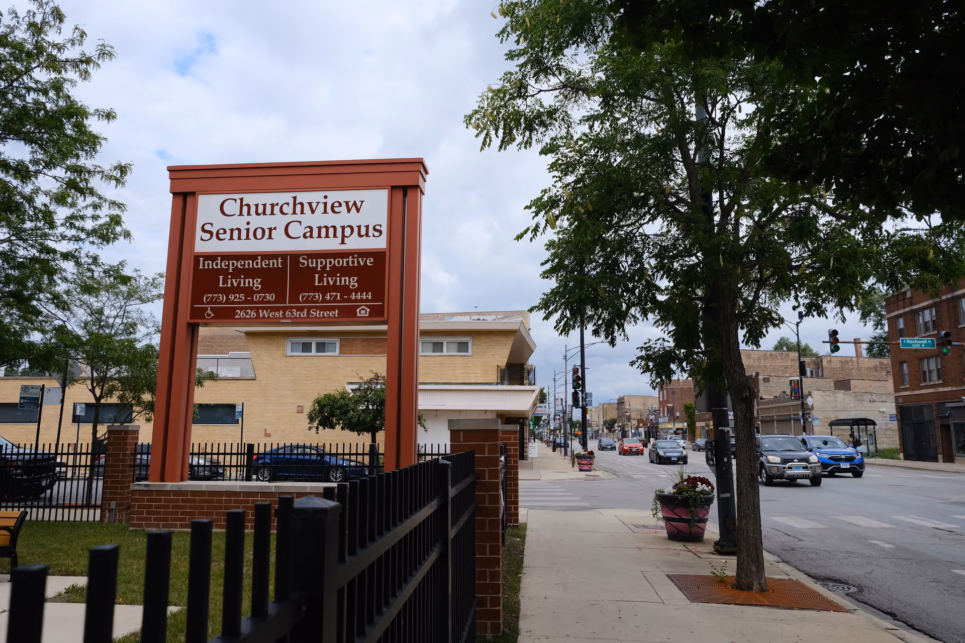 Street view showing the entrance sign for Churchview Senior Campus, advertising Independent Living and Supportive Living with contact numbers and address. The sign is mounted on a brick base with a black metal fence in front. Trees and a sidewalk line the street, with cars driving on the road and buildings in the background under a cloudy sky.