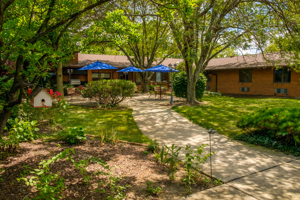 Outdoor garden area with a paved walkway leading to a patio with tables and blue umbrellas. The patio is surrounded by trees, bushes, and green grass, adjacent to a single-story brick building.