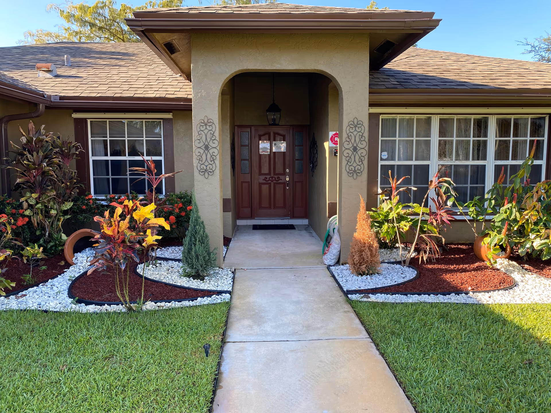 Front entrance of a single-story building with a brown door under a covered porch. The walkway leading to the door is flanked by landscaped garden beds with green grass, white rocks, red mulch, and various plants and shrubs. The building has beige walls and windows with white frames and brown shutters.