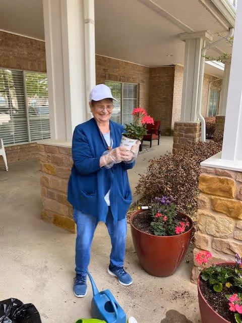 An elderly woman wearing a white cap, blue cardigan, and gloves is standing on a covered patio holding a potted pink flower. She is smiling and standing next to large red planters with various flowers and plants. The background shows a brick wall and windows of a building.