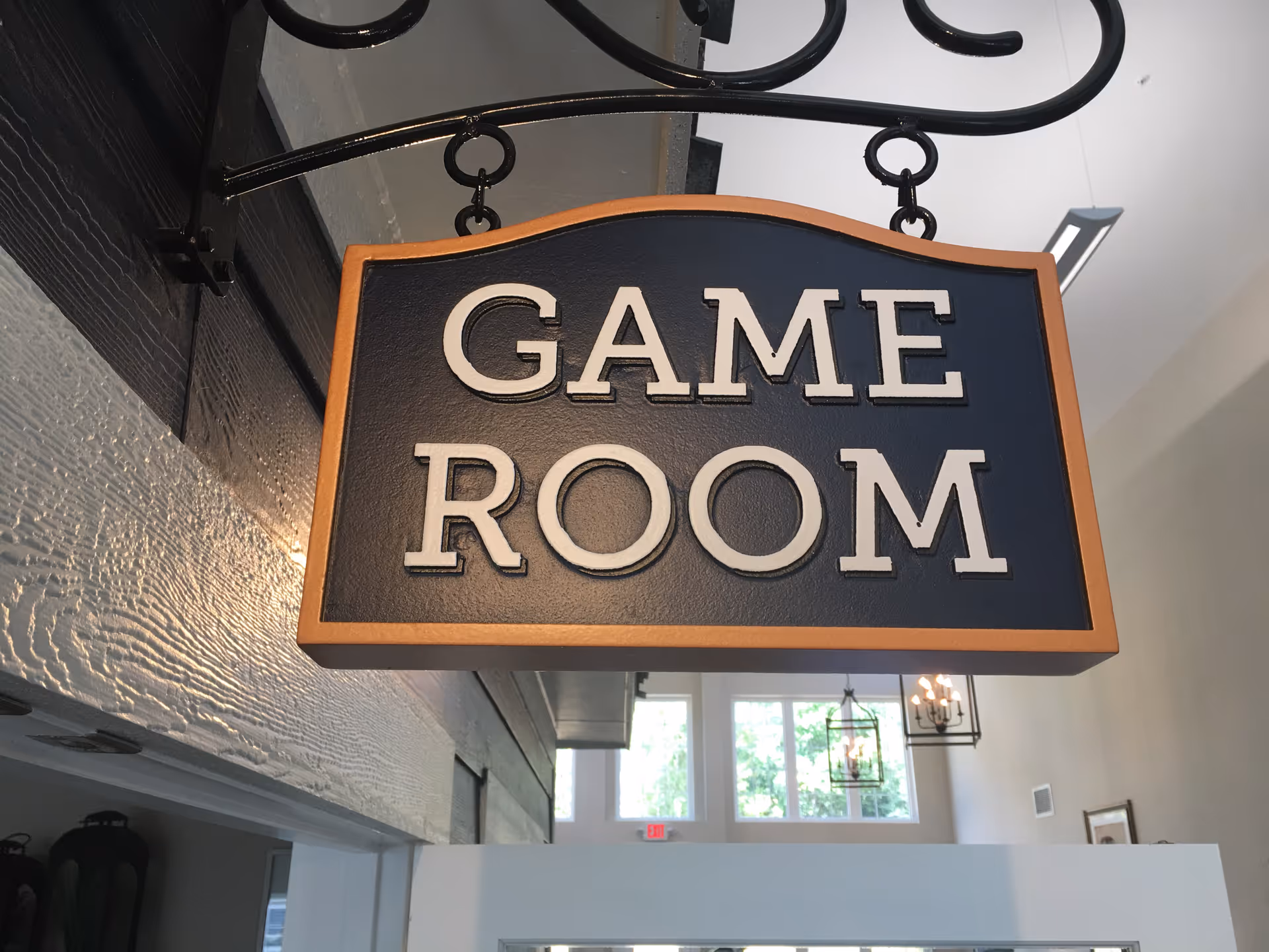 A hanging sign with a wooden frame and black background displaying the words 'GAME ROOM' in white letters, inside a building with visible windows and light fixtures in the background.