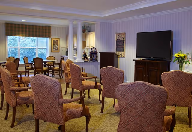 A cozy common area in a senior living facility with multiple upholstered armchairs arranged facing a large flat-screen TV on a wooden cabinet. The room has patterned carpet, wallpapered walls, and a window with a plaid valance letting in natural light. There is a small kitchen or serving area with white cabinets and decorative items, including a vase with yellow flowers on a side table.