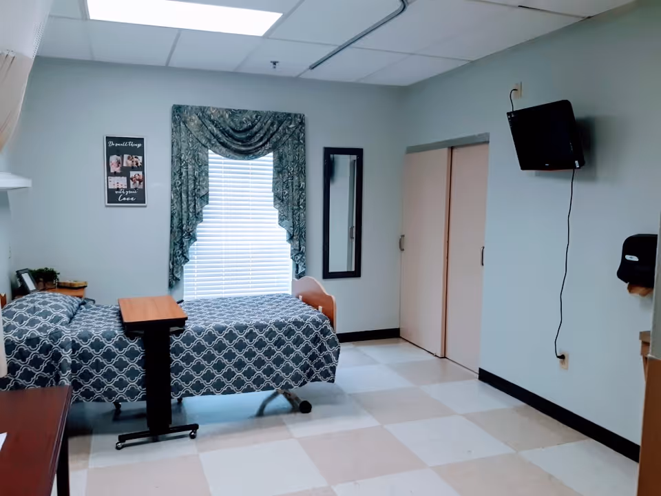 A simple, clean bedroom in a senior living facility with a single bed covered in a blue patterned bedspread. There is a small overbed table on wheels positioned over the bed. A window with blinds and decorative green curtains is centered on the back wall. To the right of the window is a full-length mirror mounted on the wall. A flat-screen TV is mounted high on the right wall with a visible power cord. The room has a tiled floor with a checkerboard pattern and light-colored walls. A small framed picture hangs on the left wall above the bed, and a paper towel dispenser is mounted on the right wall near the TV.