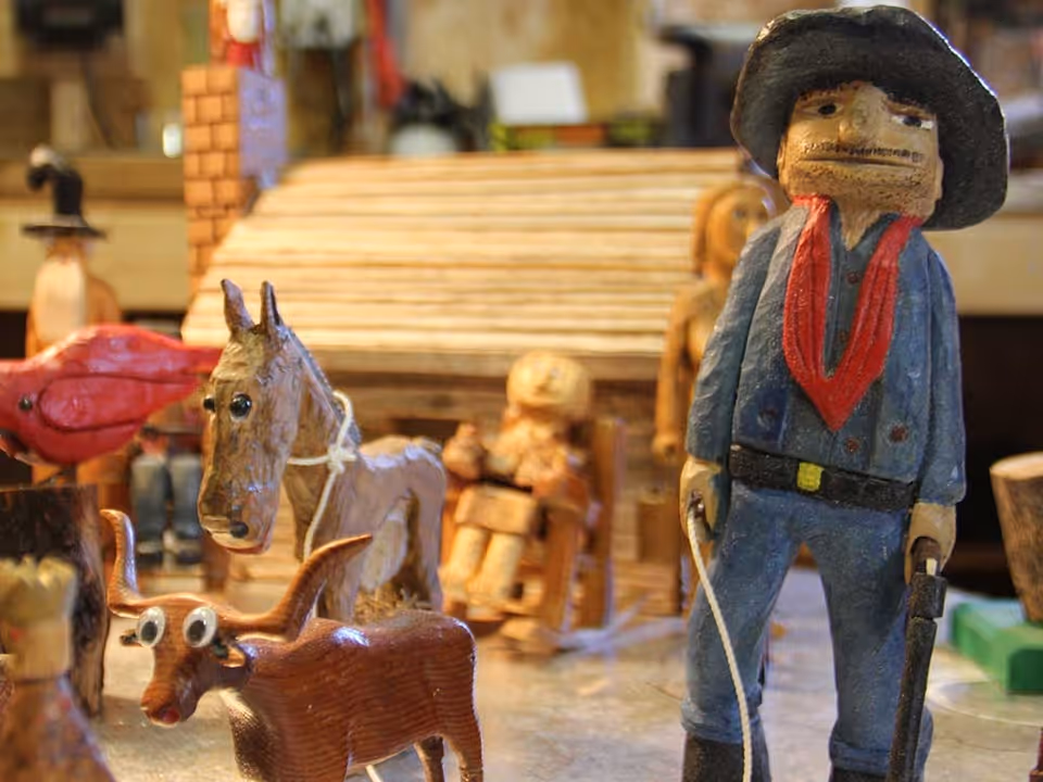A close-up view of various handcrafted wooden figurines including a cowboy wearing a hat and red scarf, a horse, a longhorn steer with googly eyes, and other small wooden characters and structures in the background.