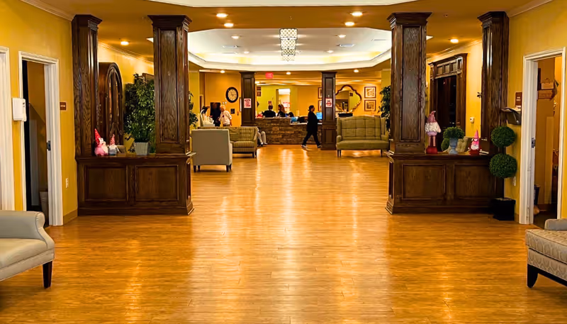 Spacious interior hallway of a senior living facility with wooden flooring, yellow walls, and dark wooden columns. Several cushioned chairs and sofas are arranged along the sides. People are visible in the background near a reception desk.