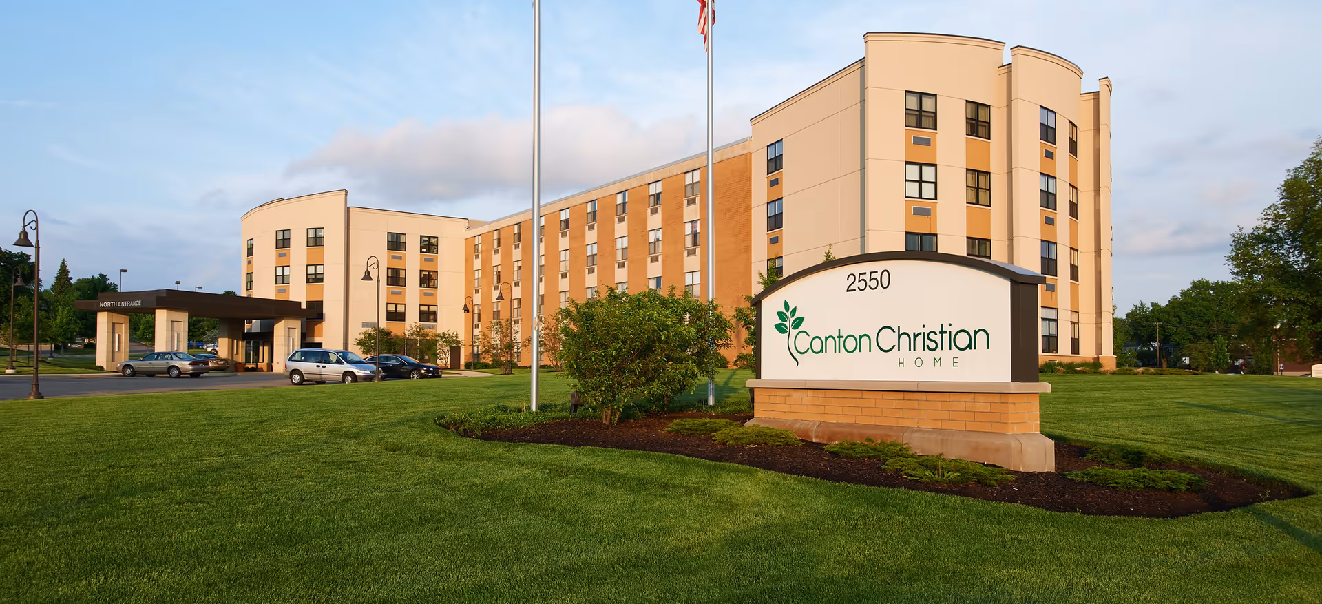Exterior view of Canton Christian Home, a multi-story senior living facility with a large green lawn in front, a sign displaying the facility name and address 2550, and several cars parked near the north entrance under a covered driveway.