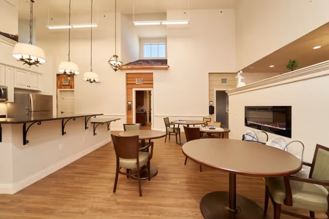 A bright and spacious common area with several round tables and chairs arranged on a wooden floor. The room features a modern electric fireplace built into a partial wall, pendant lights hanging from the ceiling, and a kitchen area with a countertop and stainless steel appliances in the background. The walls are painted in neutral tones, and there is a small window above a doorway with wood paneling around it.