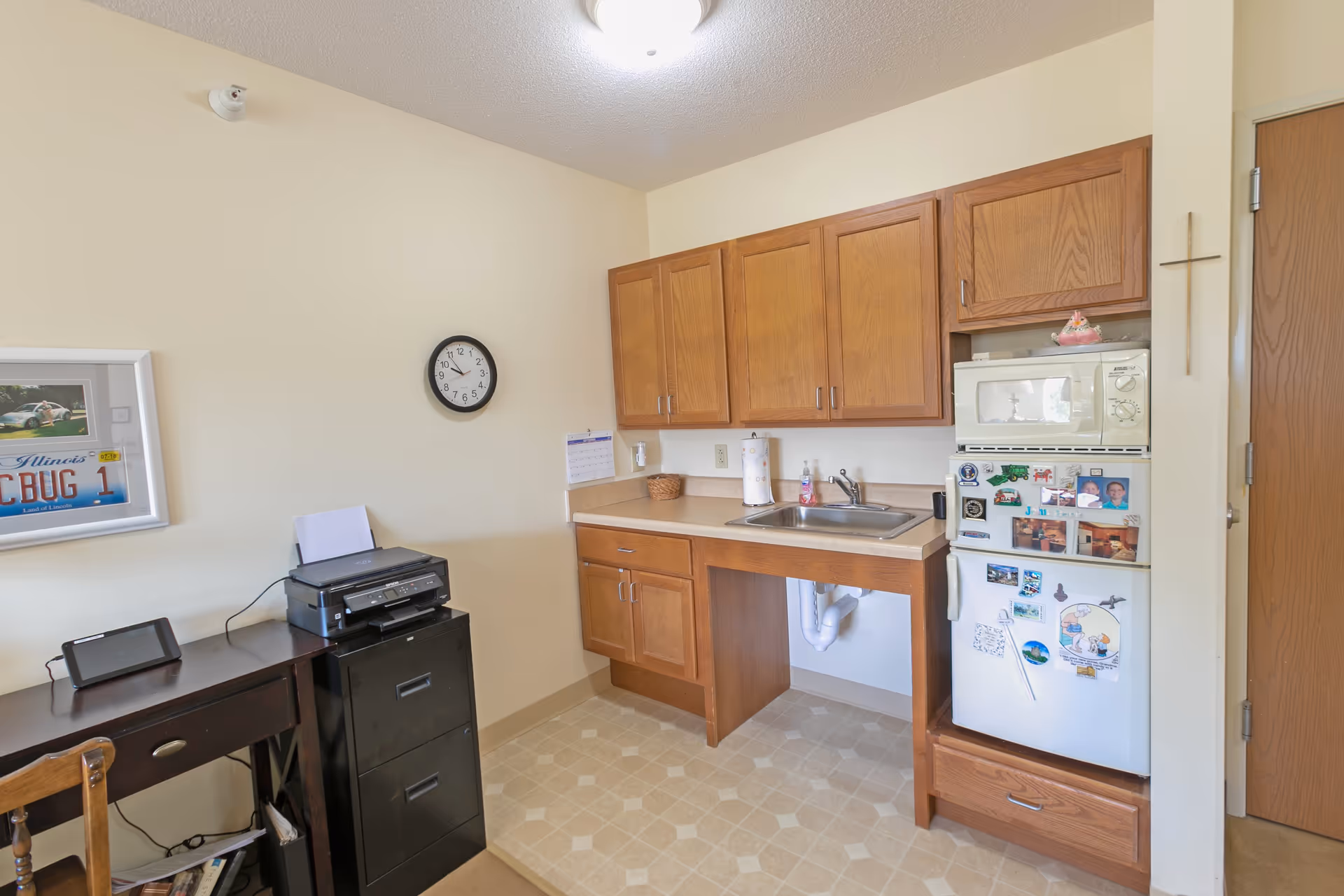 Small kitchenette area with wooden cabinets, a sink, a microwave on top of a refrigerator covered with magnets and photos, and a wooden desk with a printer and a tablet. A clock is mounted on the wall above the desk, and a framed picture with a license plate is also visible on the wall.