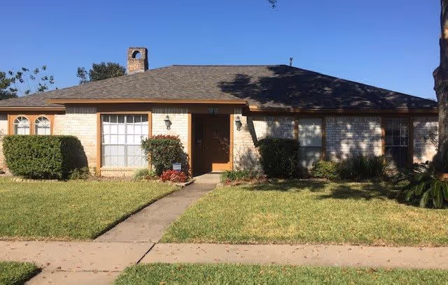 Single-story brick house with a shingled roof, a chimney, and multiple windows. The front yard has a well-maintained lawn, bushes, and a concrete walkway leading to the front door. The sky is clear and blue.