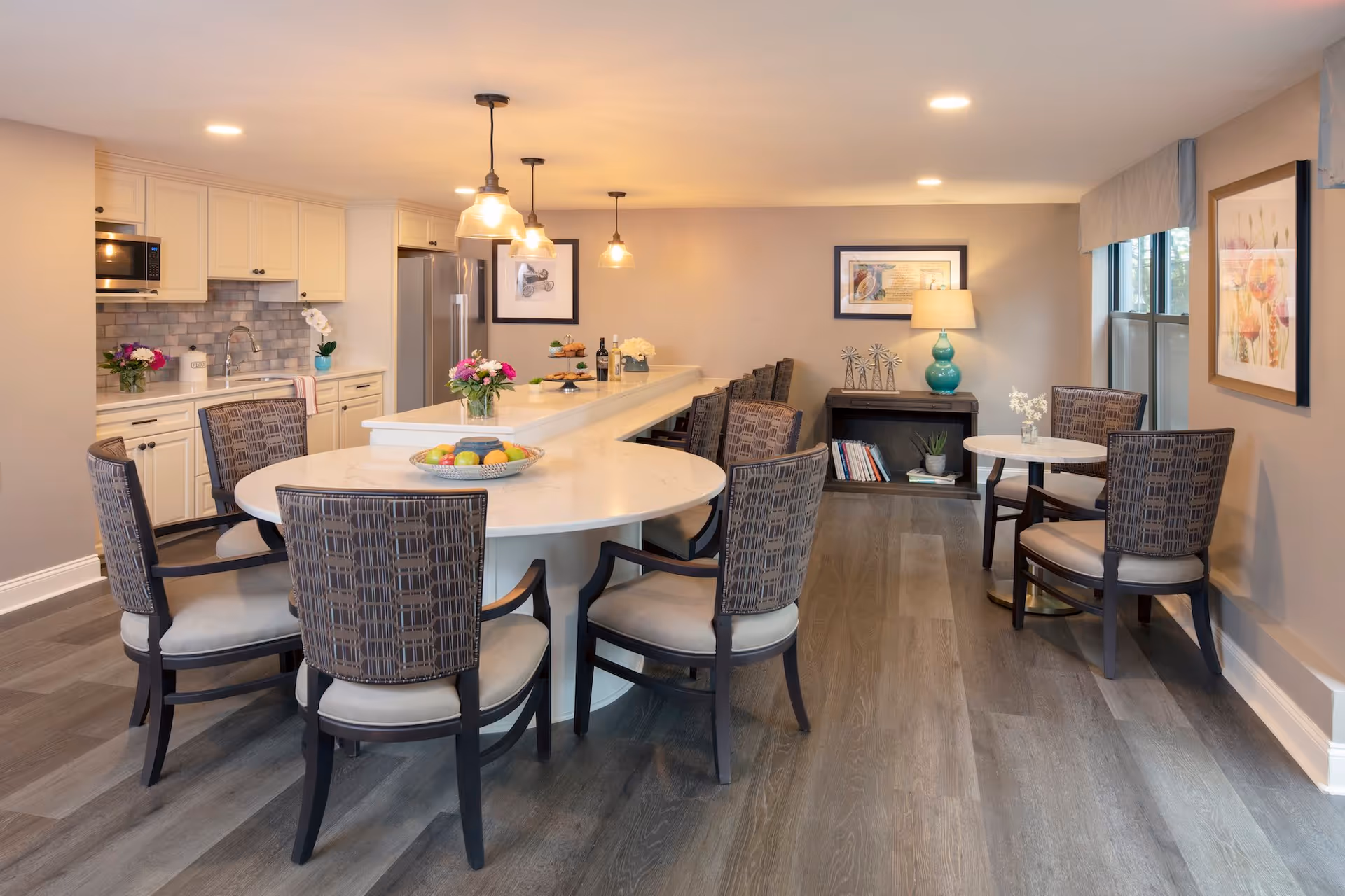 A bright and modern dining area with a large white marble countertop island surrounded by cushioned chairs. The kitchen area in the background features white cabinets, a stainless steel refrigerator, and a microwave. There are pendant lights hanging above the island, and the room is decorated with flowers, framed artwork, and a small bookshelf with decorative items and a lamp.