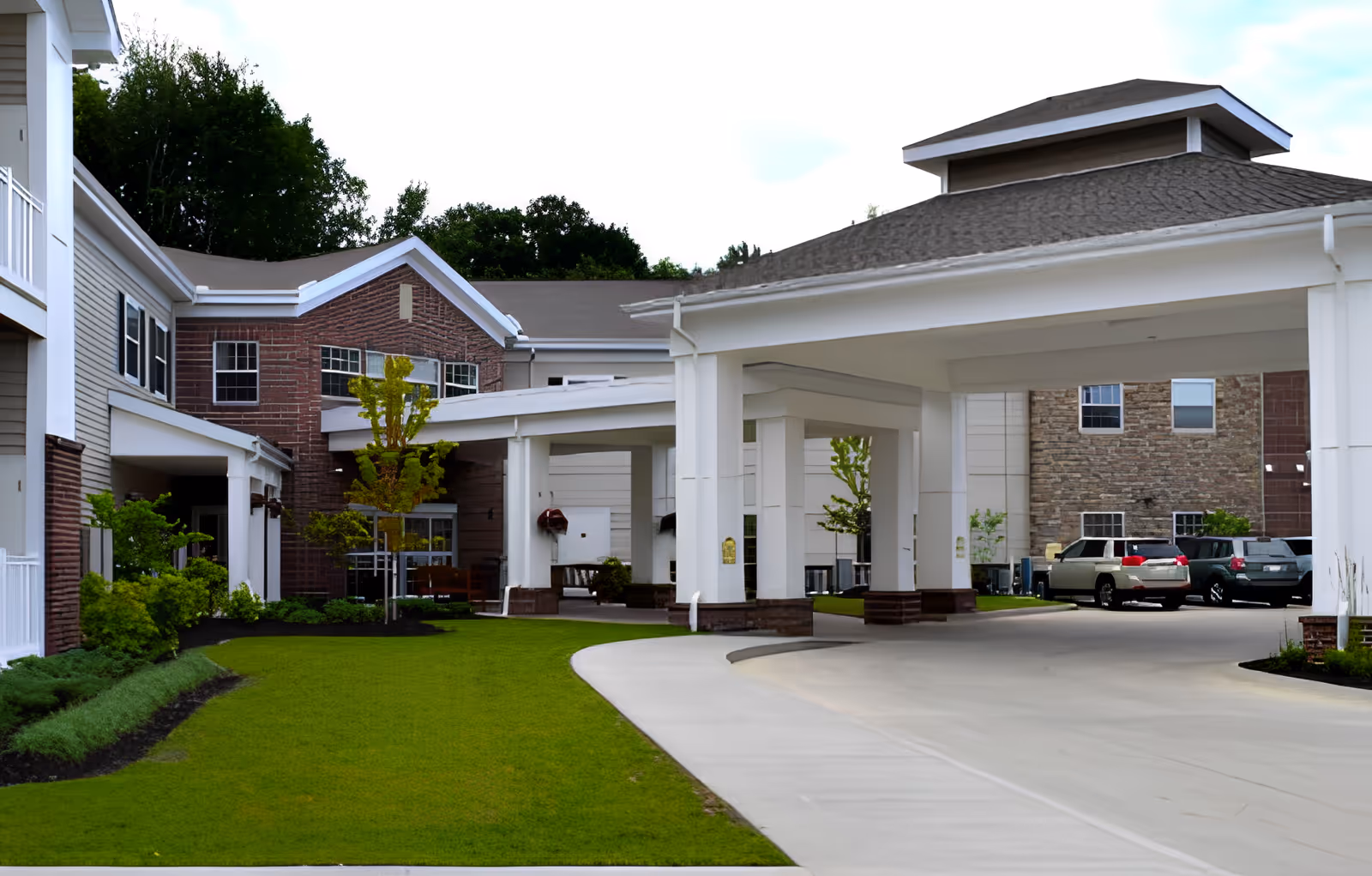 Exterior view of Danbury Mentor senior living facility showing a covered driveway entrance with white pillars, well-maintained green lawn, shrubs, and trees. The building features a mix of brick and siding with multiple windows and a parking area with several cars in the background.
