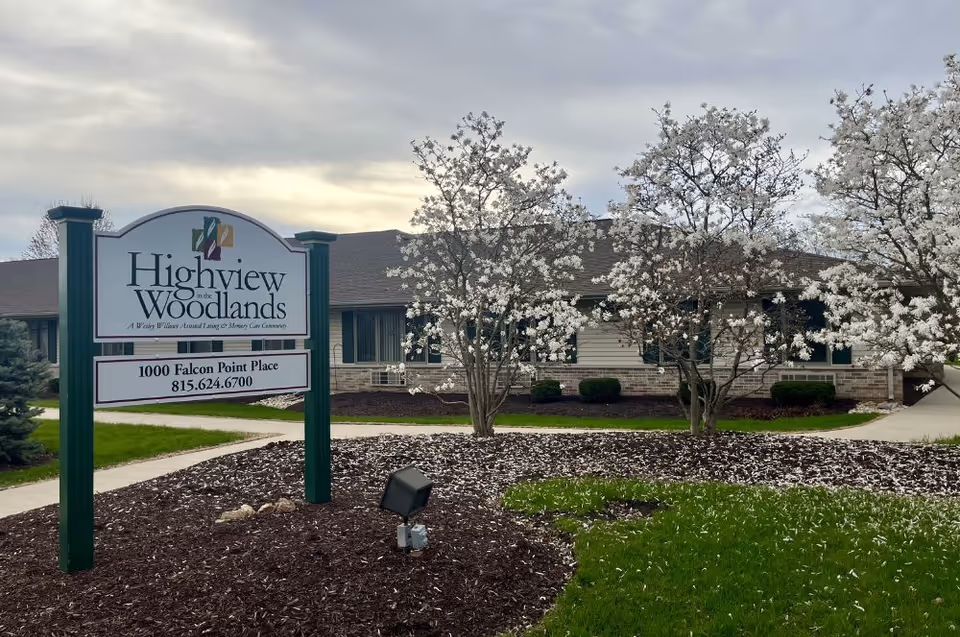 Exterior view of Highview In the Woodlands facility with a sign displaying the name and address, surrounded by blooming white flowering trees and a well-maintained lawn under a cloudy sky.