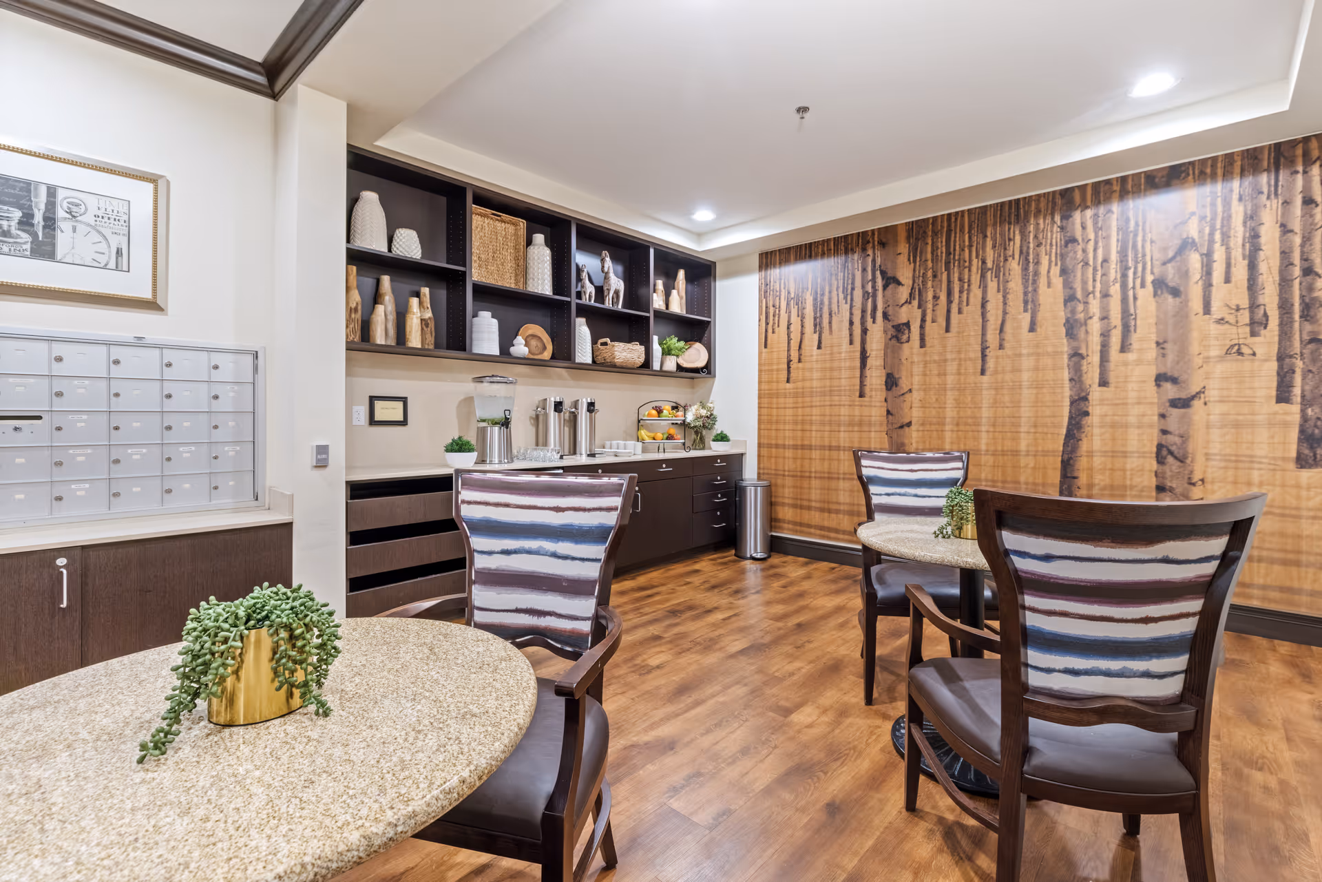 A cozy interior room with wooden flooring featuring two round tables with patterned chairs. The back wall has a large mural of a birch tree forest. On the left side, there are shelves with decorative items and a counter with a water dispenser, coffee urns, and fruit basket. A set of mailboxes is mounted on the wall near the corner.