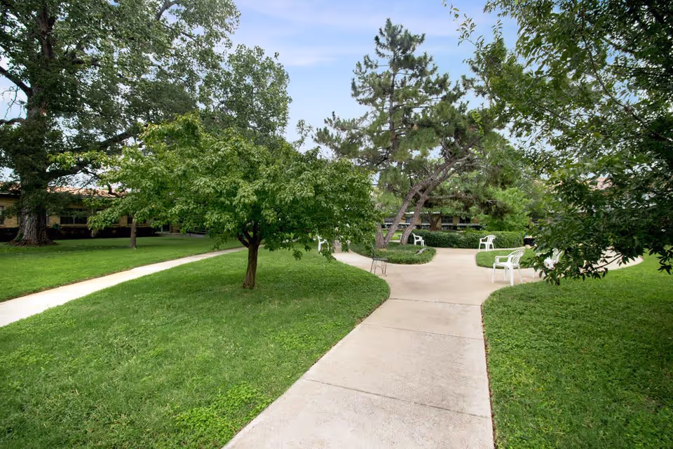 A landscaped outdoor area with green grass, several trees, and a concrete walkway that splits into two paths. White plastic chairs are placed along the walkway, and a building is partially visible in the background under a partly cloudy sky.