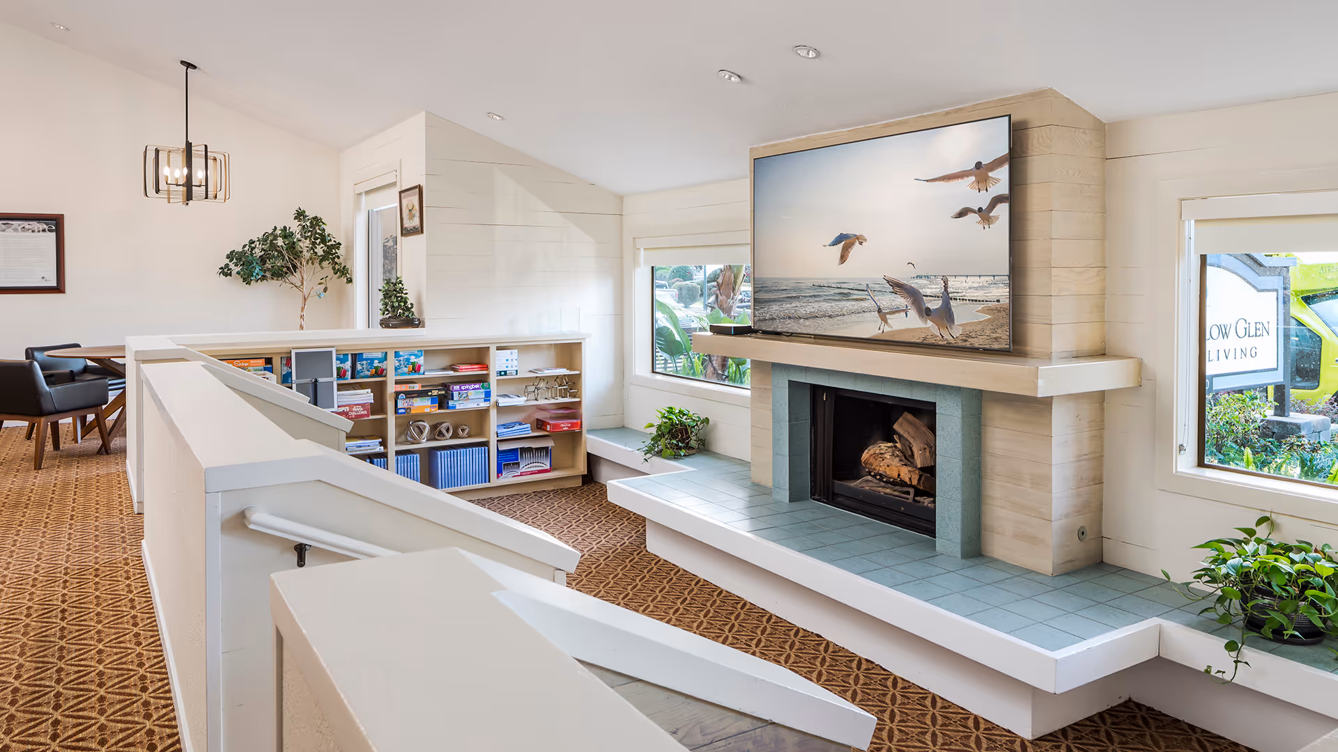A bright and cozy common area with a fireplace and a large TV mounted above it displaying an image of seagulls flying over the ocean. The room features a patterned carpet, built-in shelves with board games and books, a small seating nook by the window with plants, and a dining area with chairs and a hanging light fixture in the background.