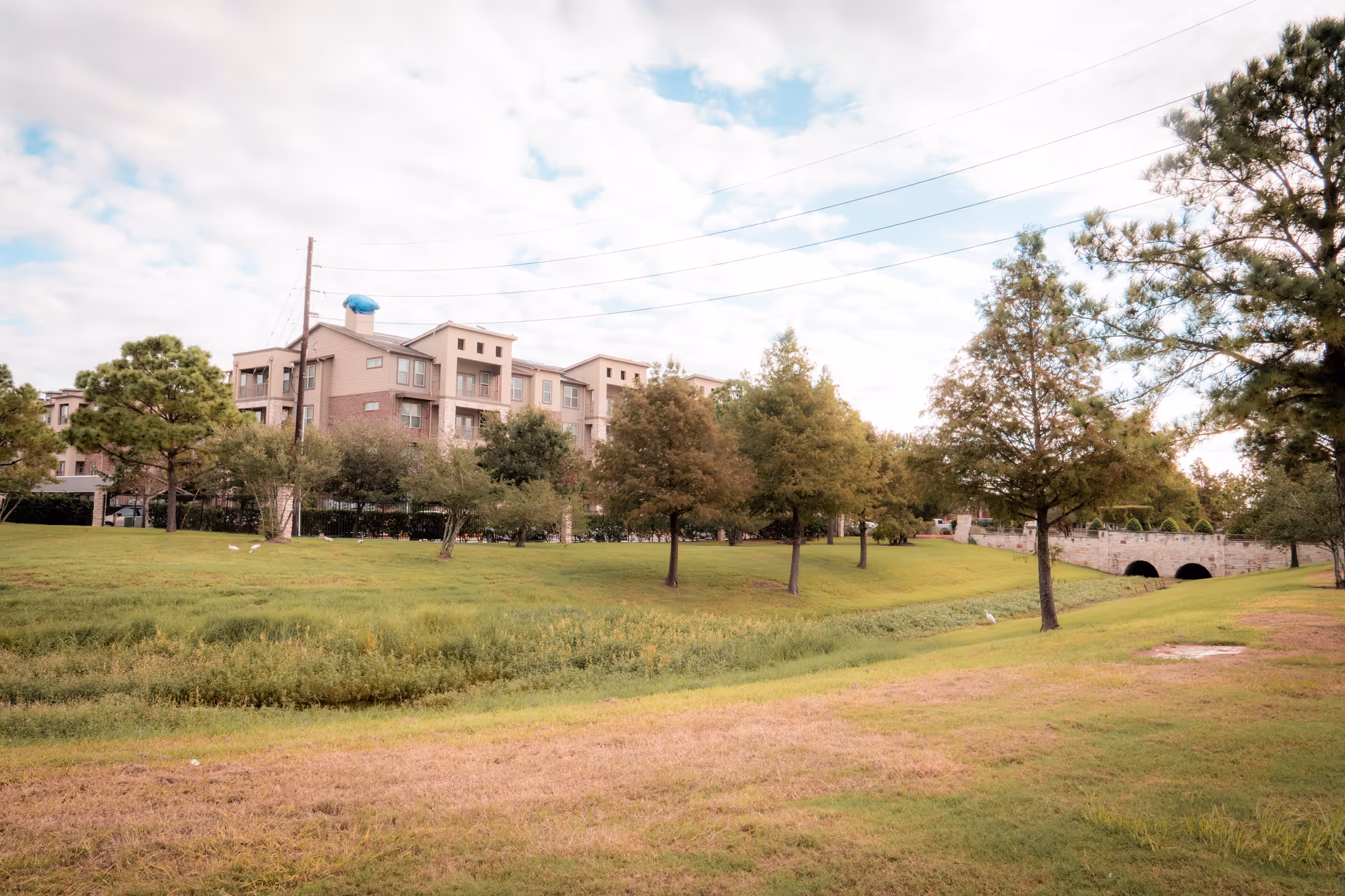 A landscaped outdoor area with green grass, several trees, and a small creek or drainage ditch. In the background, there is a multi-story residential building with balconies. The sky is partly cloudy.