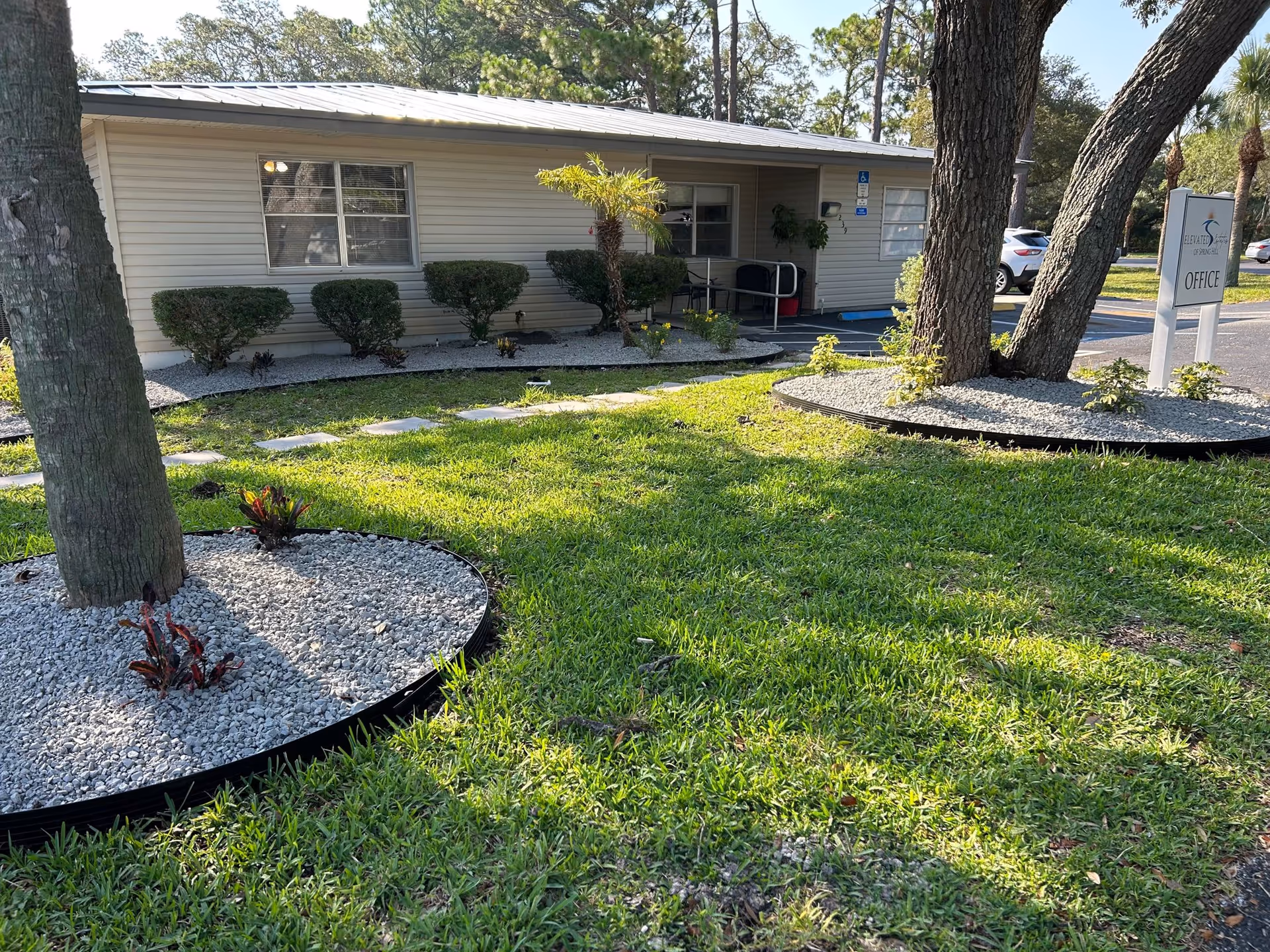 Exterior view of a single-story building with beige siding, surrounded by green grass and landscaped areas with white gravel and small plants. There are two large trees in the foreground with circular gravel beds around their bases. A sign near the building reads 'Office'.