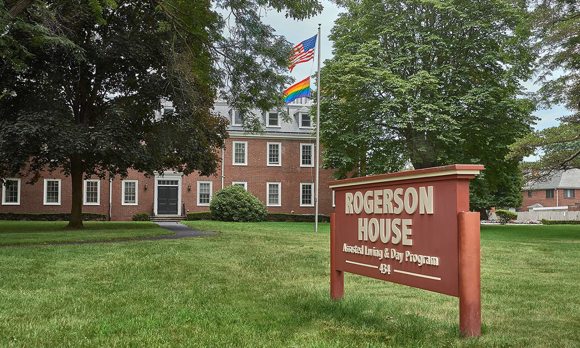 Exterior view of Rogerson House Assisted Living & Day Program building with a large green lawn, trees, and two flagpoles flying the American flag and a rainbow pride flag.