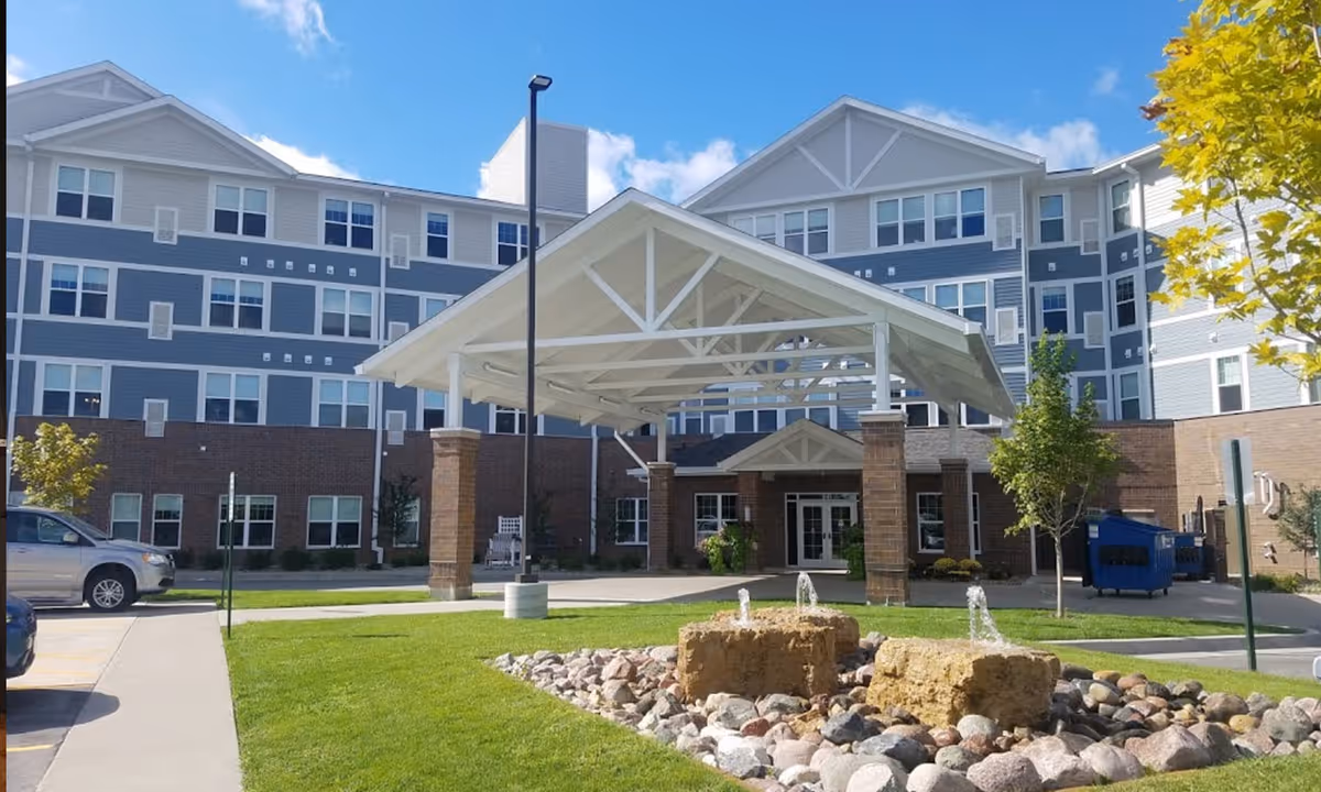 Front exterior view of Overlook Village facility showing a multi-story building with a covered entrance supported by brick pillars, a small landscaped area with rocks and water fountains, green grass, trees, and a clear blue sky.