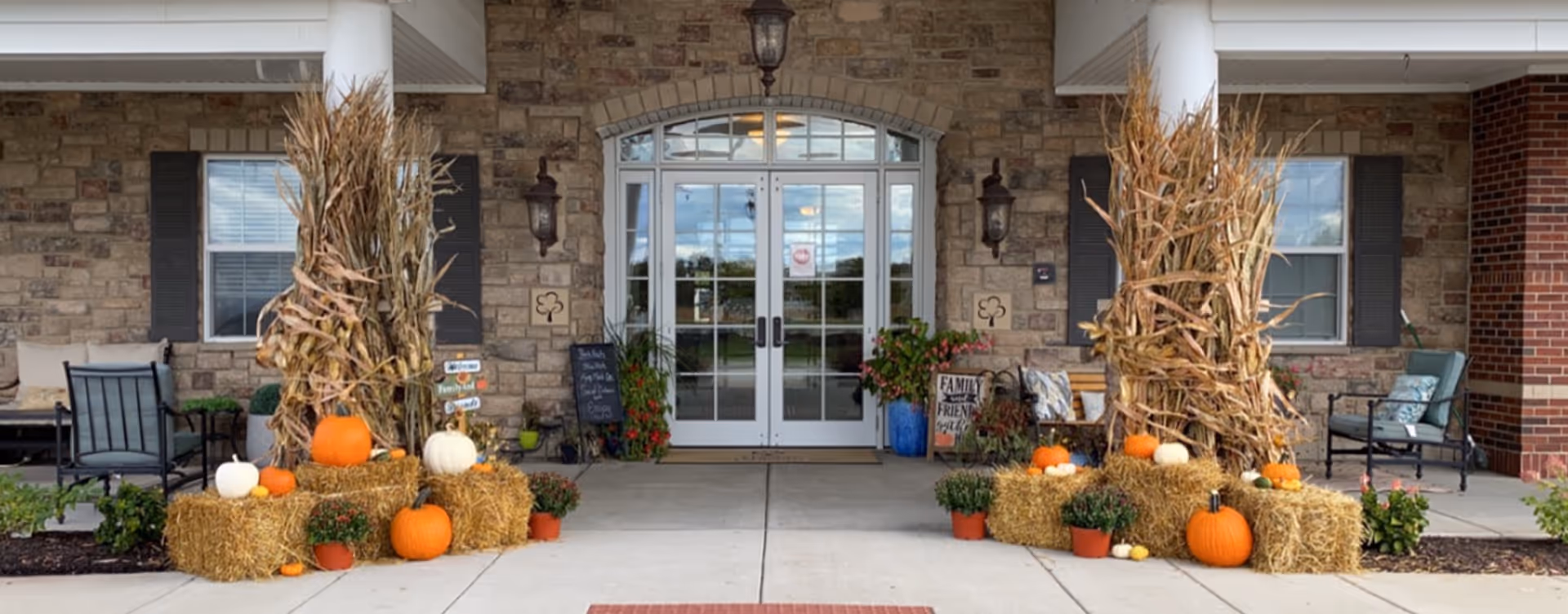 Entrance of a building with double glass doors framed by stone walls and white columns. The entrance is decorated with hay bales, pumpkins, dried corn stalks, and potted plants, creating a fall-themed display. There are chairs and small tables on either side of the entrance.