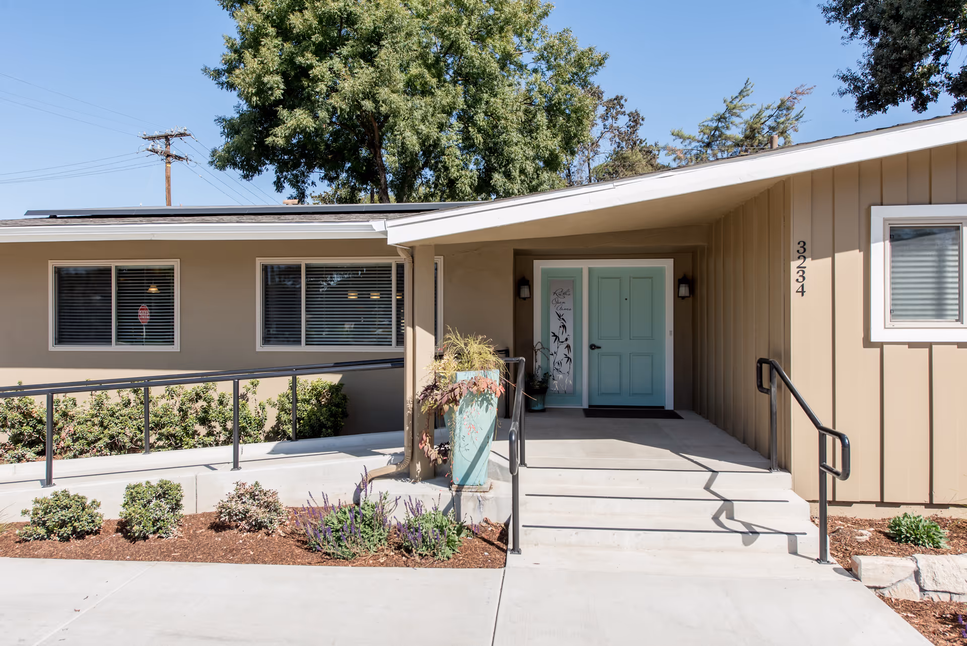 Exterior view of the entrance to Kaweah Health Ruth Wood Open Arms House, featuring a beige building with a light blue double door, a ramp with handrails, stairs, and landscaping with shrubs and flowers under a clear blue sky.