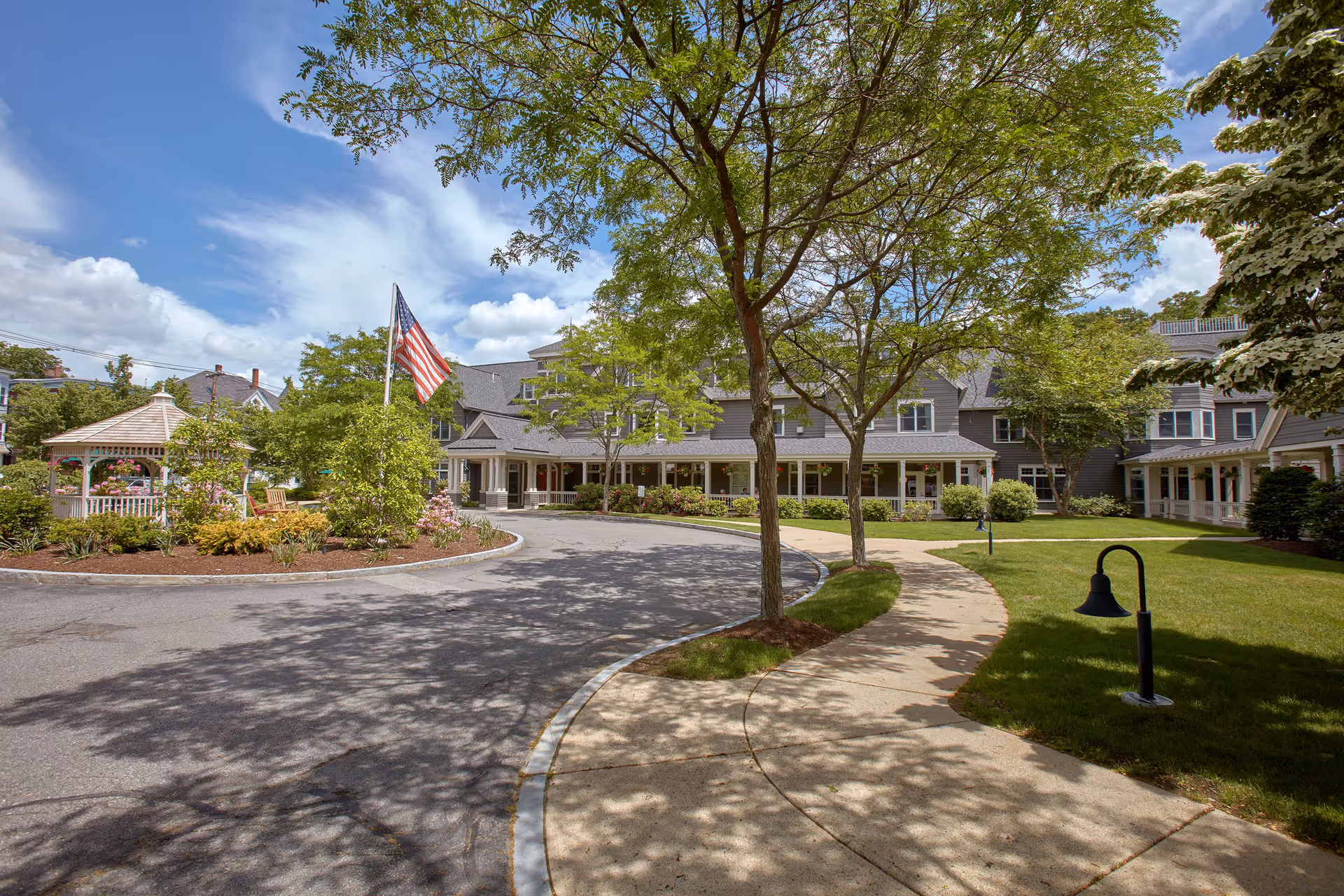 Exterior view of Goddard House Assisted Living facility showing a curved driveway, a sidewalk, trees, a gazebo, and an American flag under a partly cloudy sky.