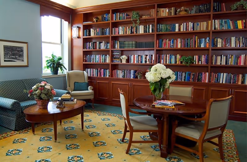 A cozy library room with a large wooden bookshelf filled with books along the back wall. In front of the bookshelf is a round wooden table with four chairs, and a vase with white flowers on the table. To the left, there is a green patterned sofa, a wooden coffee table with a flower arrangement and books, and a beige armchair near a window with natural light coming in. The floor is covered with a yellow patterned carpet.
