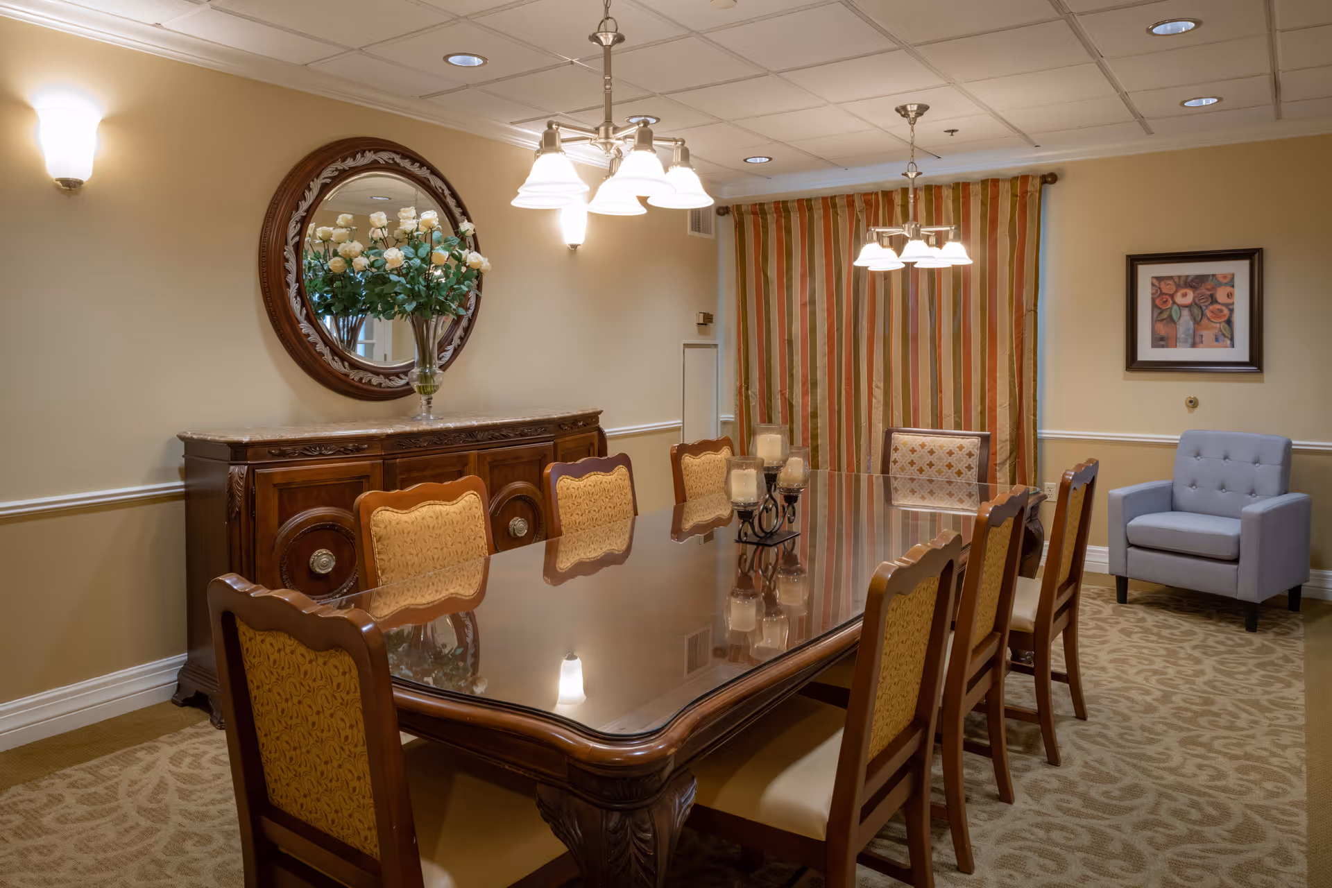 A formal dining room with a long polished wooden table surrounded by eight upholstered chairs. On the table is a decorative candle holder with multiple candles. The room features a wooden sideboard with a large round mirror above it, a vase with white flowers, striped curtains, a framed floral painting on the wall, and a gray upholstered armchair in the corner. The lighting includes ceiling recessed lights and two hanging light fixtures above the table.