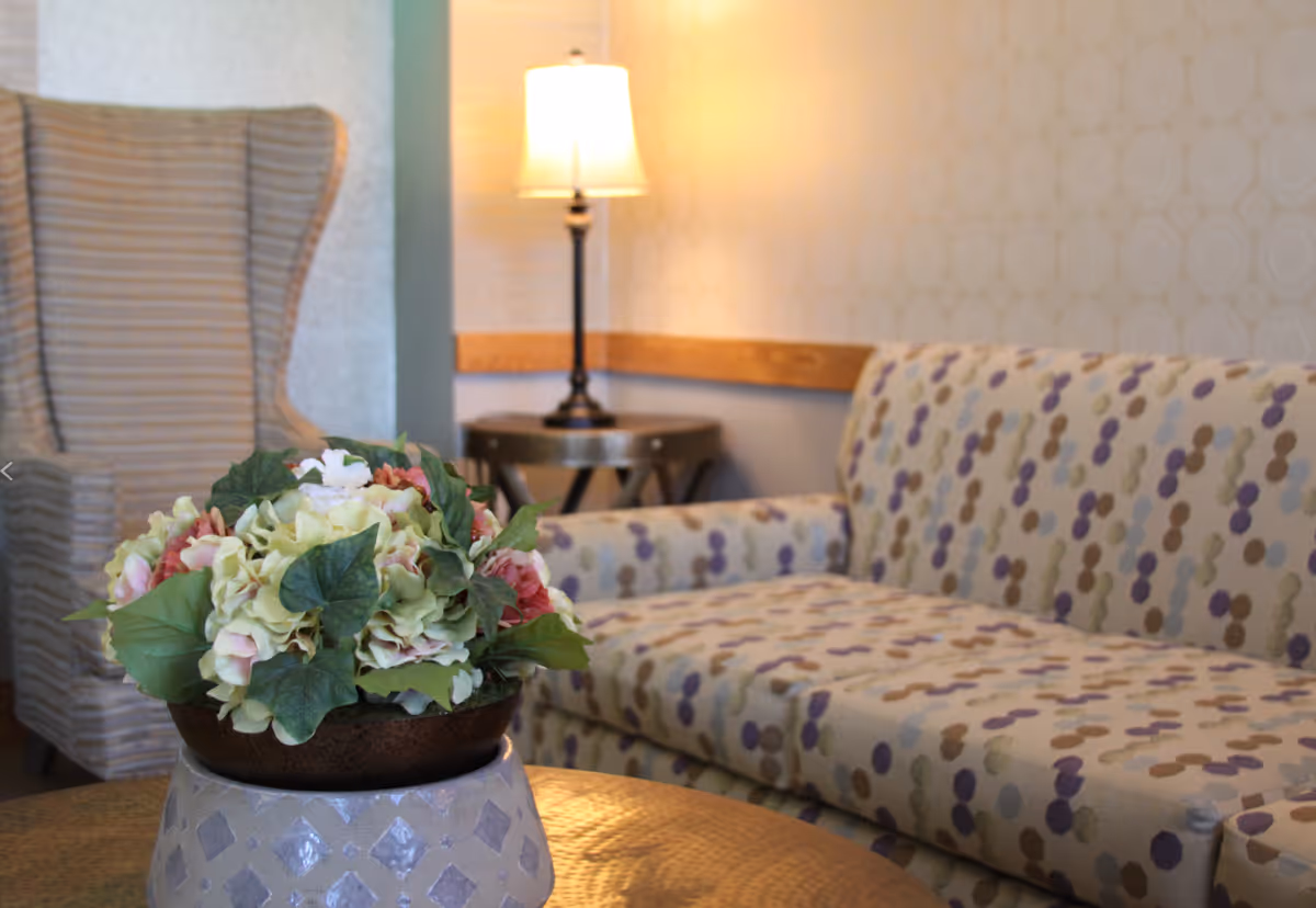 A cozy living room area with a patterned sofa, a striped wingback chair, a round table with a floral arrangement, and a lit table lamp on a small side table against a softly patterned wall.