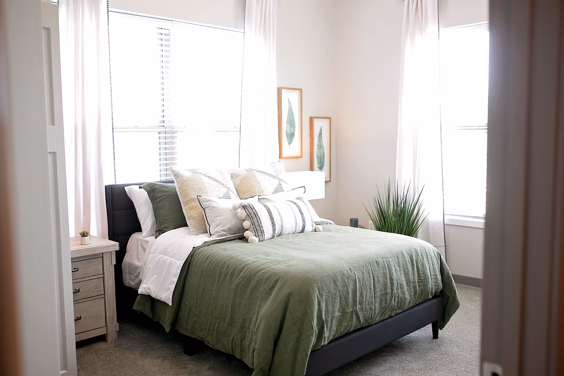 A bright bedroom with a neatly made bed featuring green bedding and multiple decorative pillows. There are two windows with white curtains allowing natural light to fill the room. A small wooden nightstand with drawers and a small plant is beside the bed. Two framed botanical prints hang on the wall above a white lamp and a green potted plant is placed near the window.