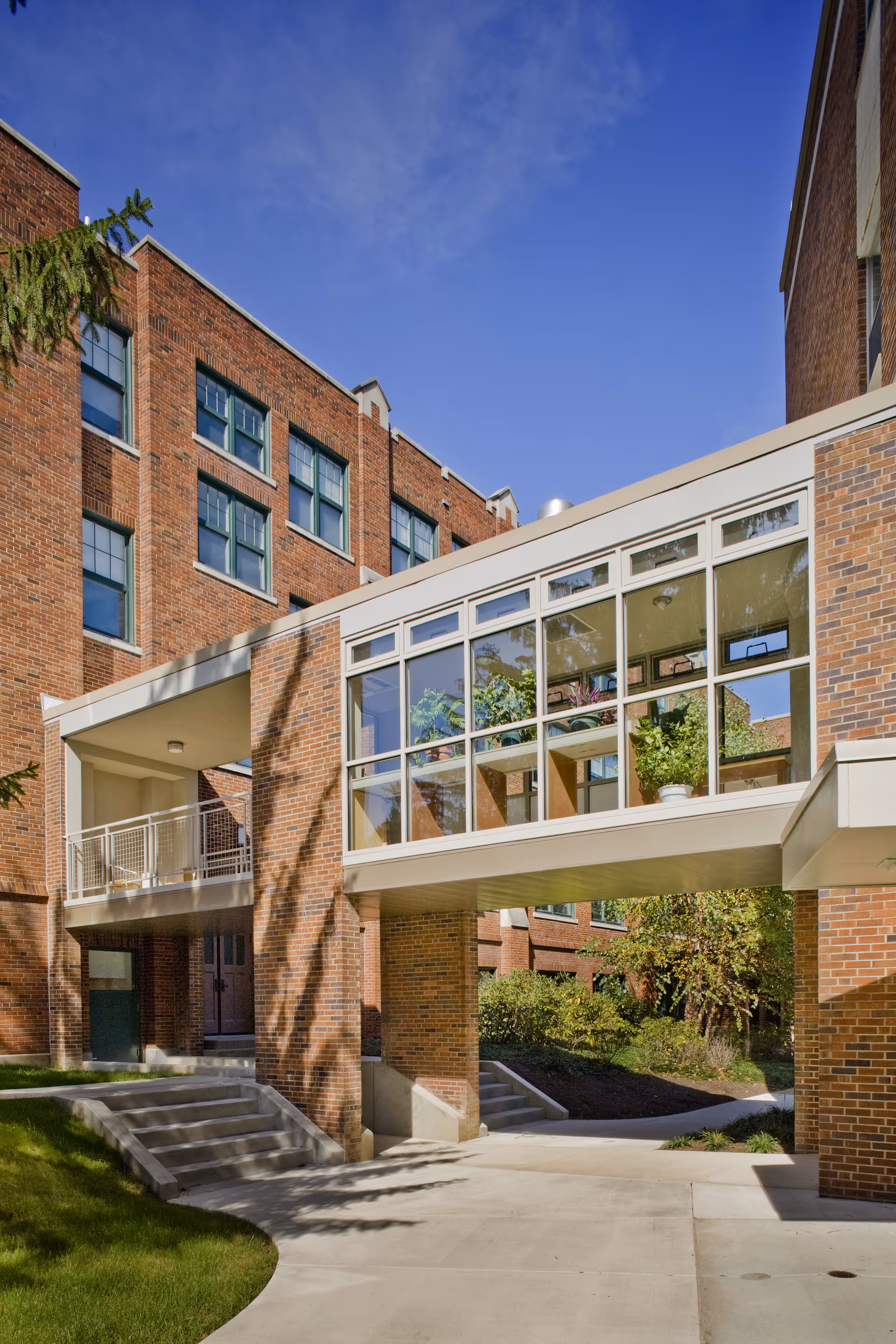 Exterior view of a brick building with a glass-enclosed walkway connecting two sections. There are stairs and a ramp leading to entrances, with greenery and trees around the building under a clear blue sky.