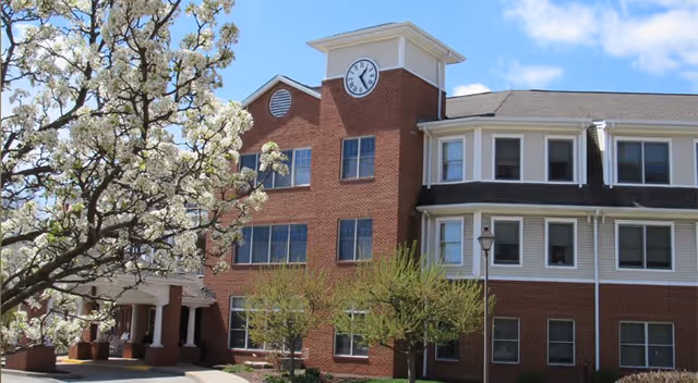 Exterior view of a multi-story brick building with white trim and a clock tower. There are blooming trees and shrubs in front of the building under a partly cloudy blue sky.