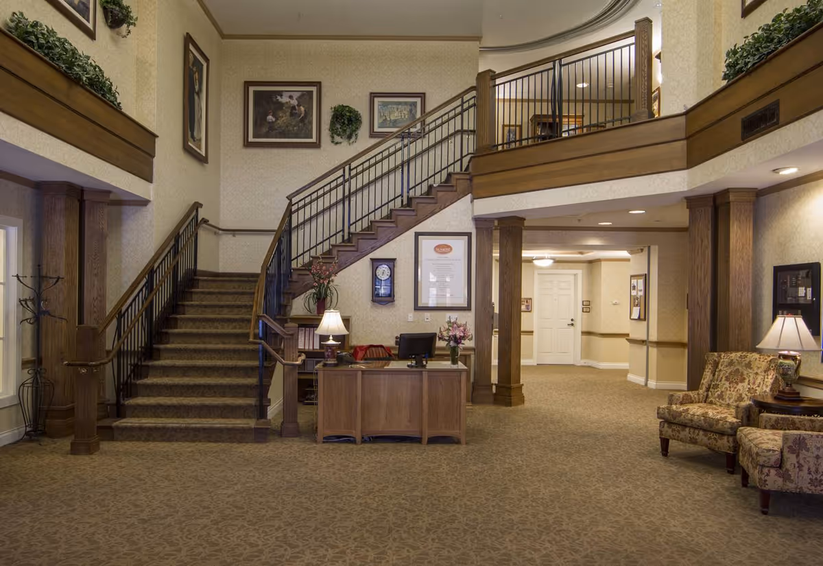 Interior view of a senior living facility lobby with a wooden reception desk, a computer, a lamp, floral arrangements, a staircase with carpeted steps and metal railings, framed artwork on the walls, and comfortable floral-patterned armchairs. The space features beige walls, carpeted floors, and wooden columns supporting an upper balcony.