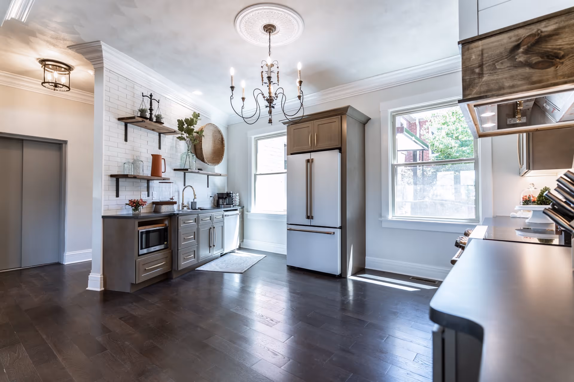 Bright modern kitchen with dark wood flooring, gray cabinets, a white refrigerator, two large windows letting in natural light, open shelves with decorative items, a chandelier hanging from the ceiling, and stainless steel appliances.